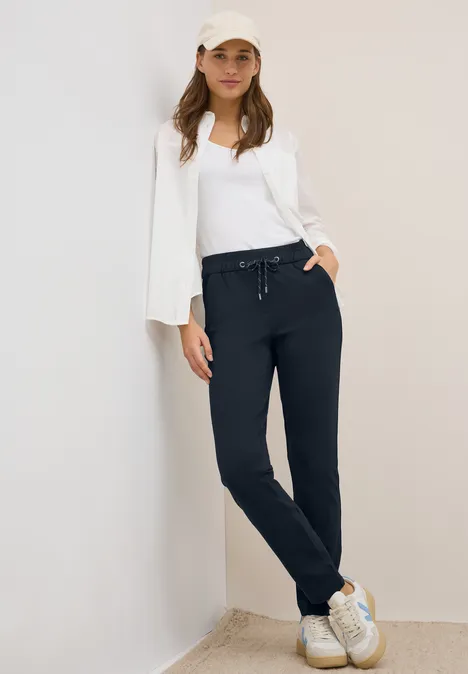 Frau in weißem Shirt, blauer Hose und Kappe lehnt an Wand.